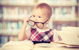 Happy funny baby girl in glasses reading a book in a library